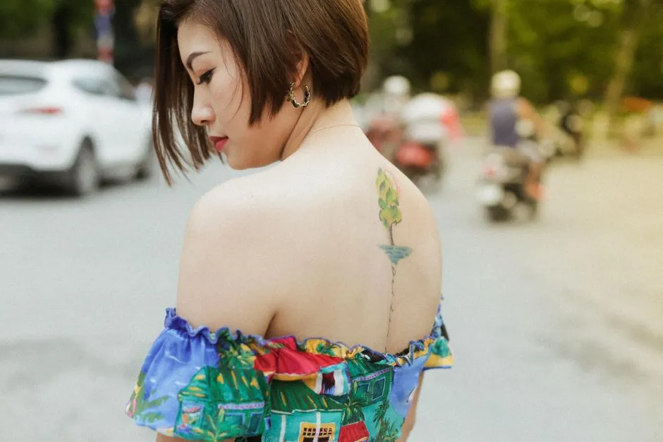 Side profile of a stylish woman with a back tattoo, standing outdoors with blurred background.